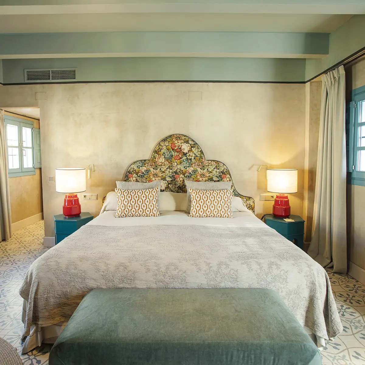 Dormitorio luminoso con cama doble, l&aacute;mparas y cabecero floral y cojines azules