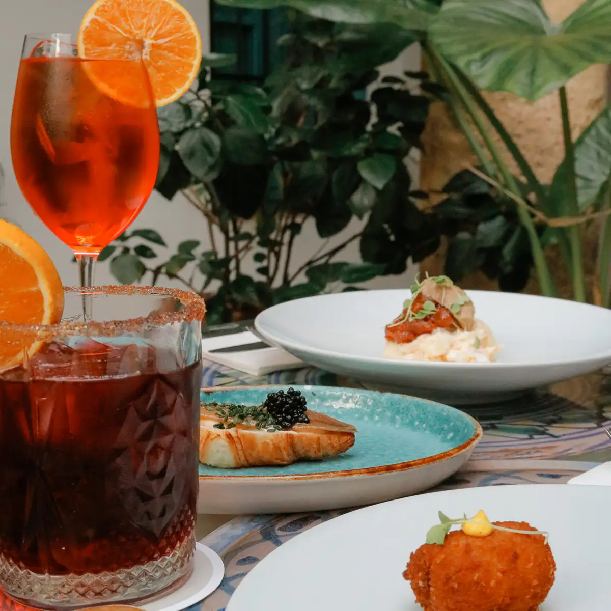 C&oacute;cteles con naranjas y platos gourmet en mesa, ambiente tropical.