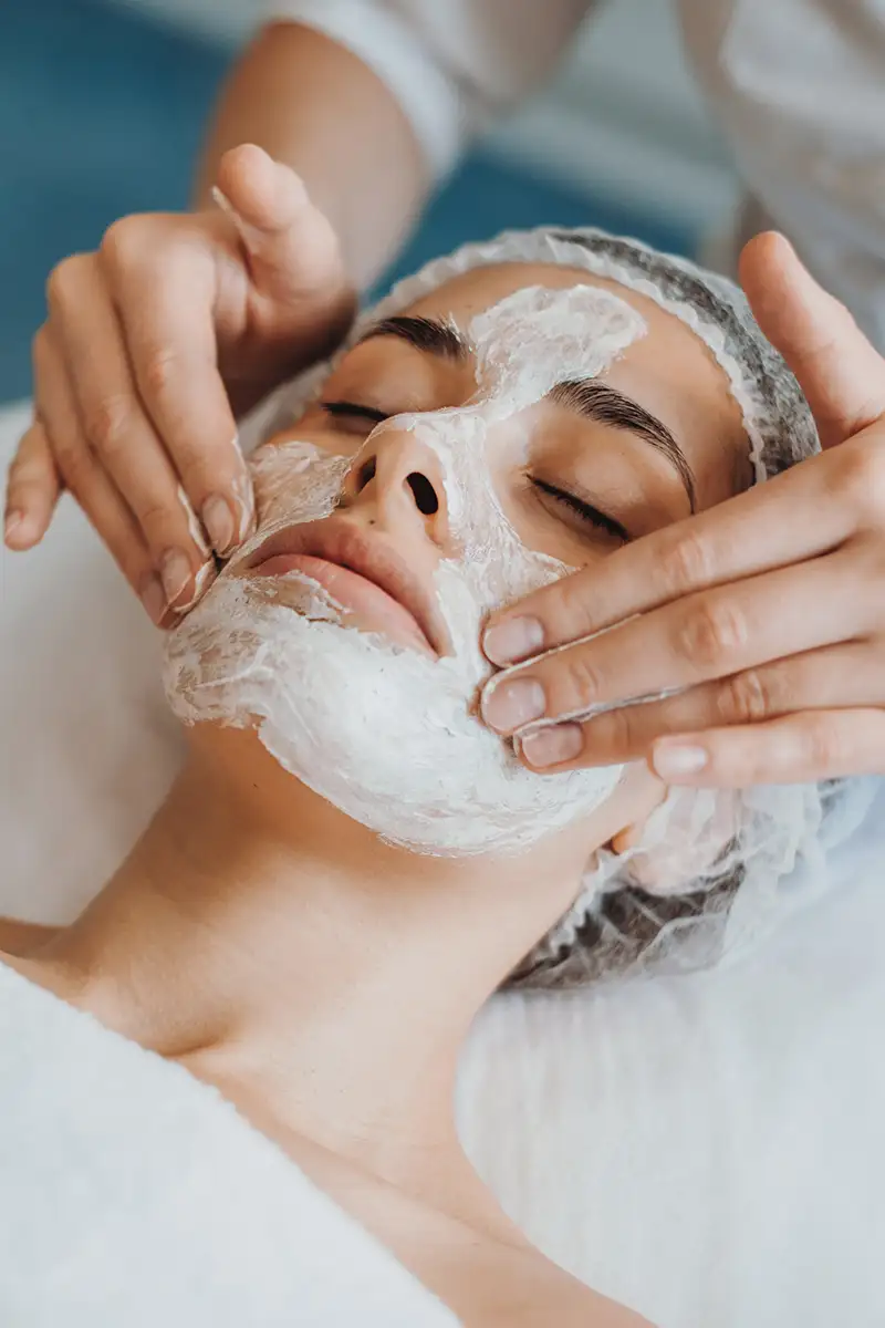 Mujer con mascarilla facial mientras una terapeuta le aplica y retira el producto