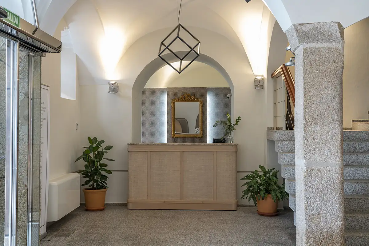 Interior de un hotel con escalera, plantas y espejo dorado bajo arco.