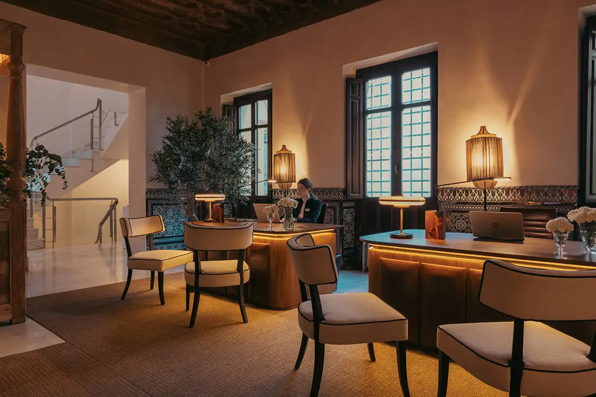 Recepci&oacute;n de hotel con recepcionista, escritorio, sillas y l&aacute;mparas de luz c&aacute;lida.