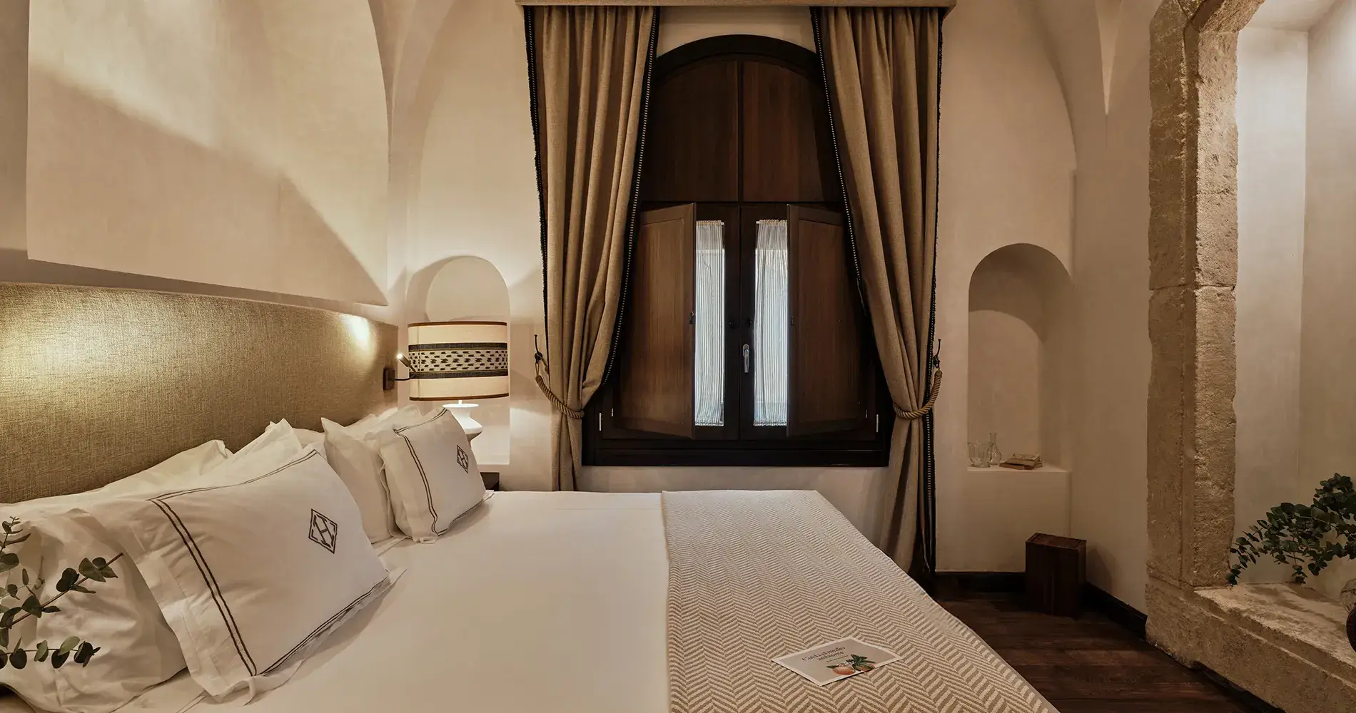 Dormitorio con cama y cortinas beige frente a puerta de madera oscura y nichos