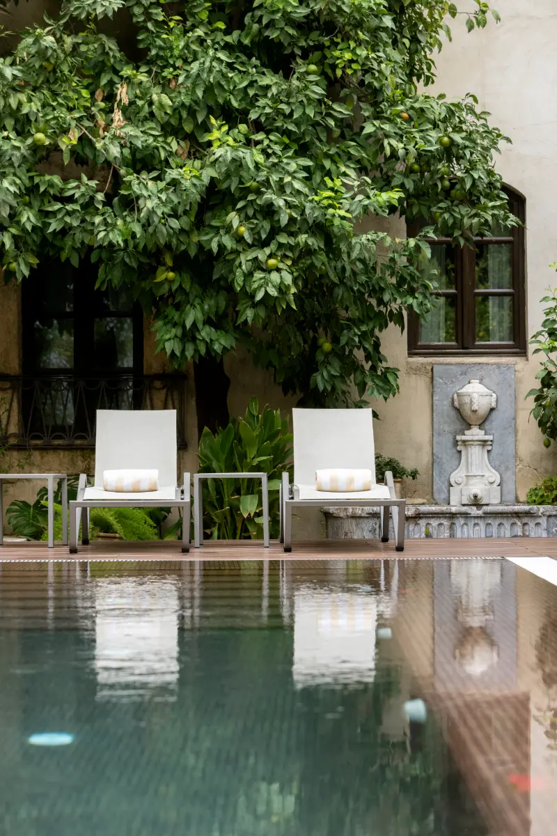 Piscina con dos tumbonas blancas bajo frondoso &aacute;rbol y fuente de pared