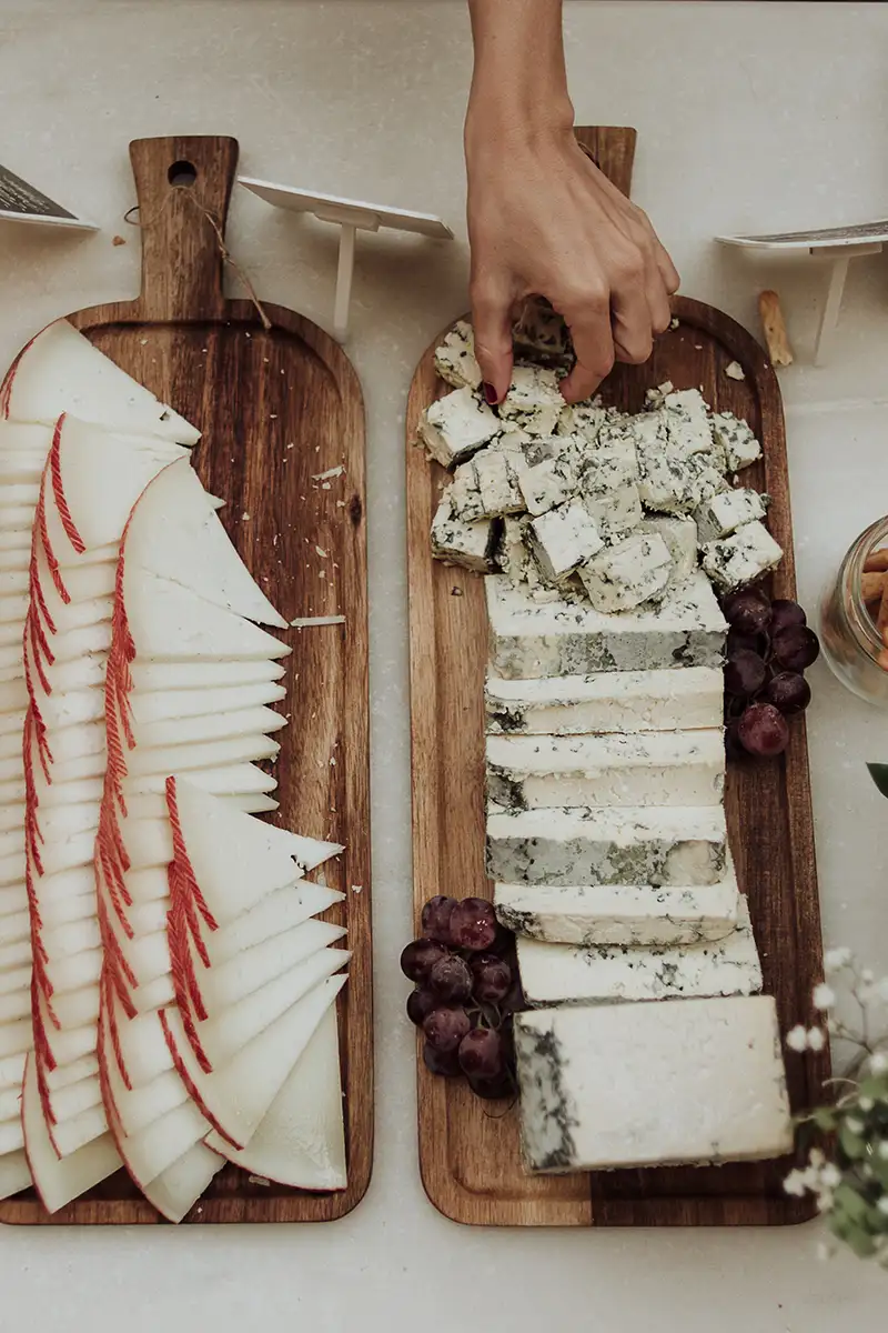 Mano desmenuza queso azul sobre tabla de madera con uvas y pan.