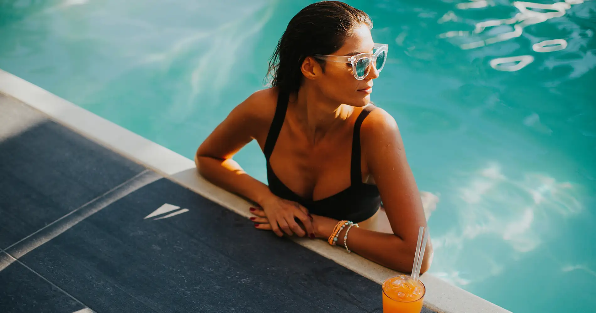 Mujer junto a la piscina con gafas, en el borde, y bebida en la mano.