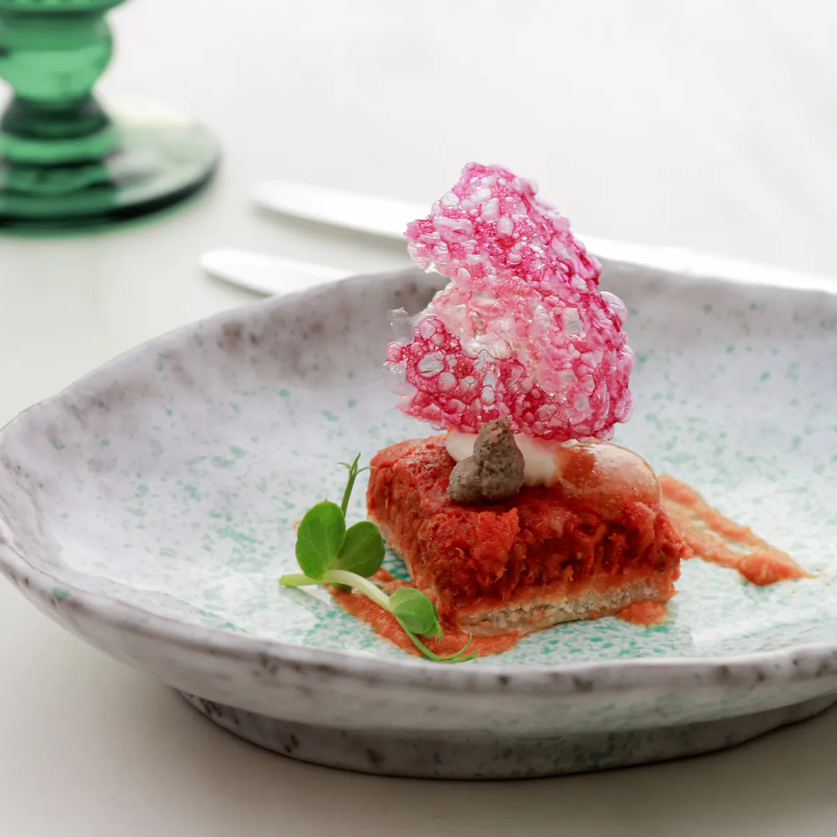 Postre con crema rosa sobre base roja, en plato decorado, con hierbas verdes