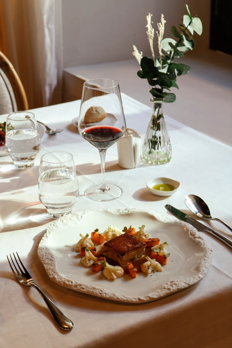 Plato de carne con verduras y vino en una mesa con mantel blanco y flores.