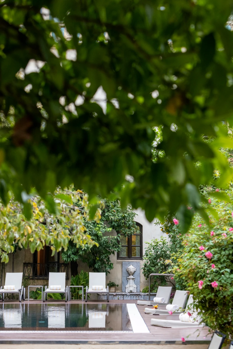 Patio con piscina y tumbonas, entre jardines verdes y flores.