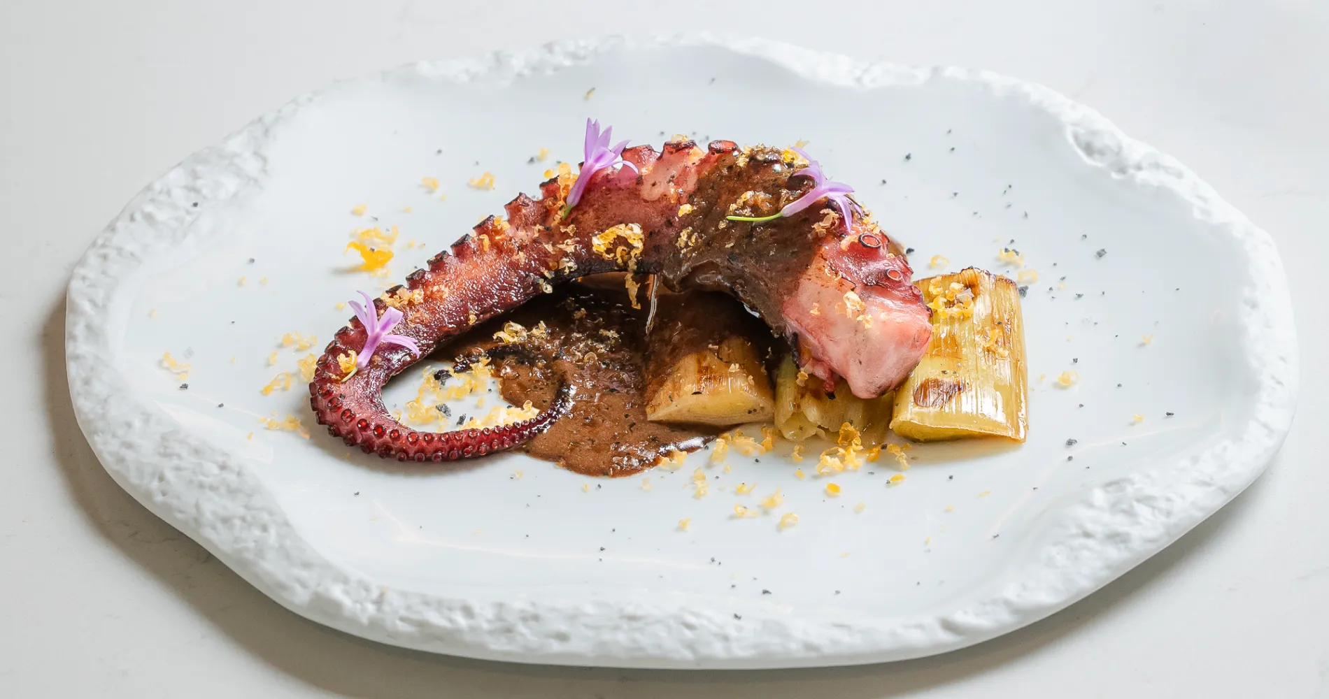 Pulpo a la parrilla sobre plato blanco con salsas y flores moradas