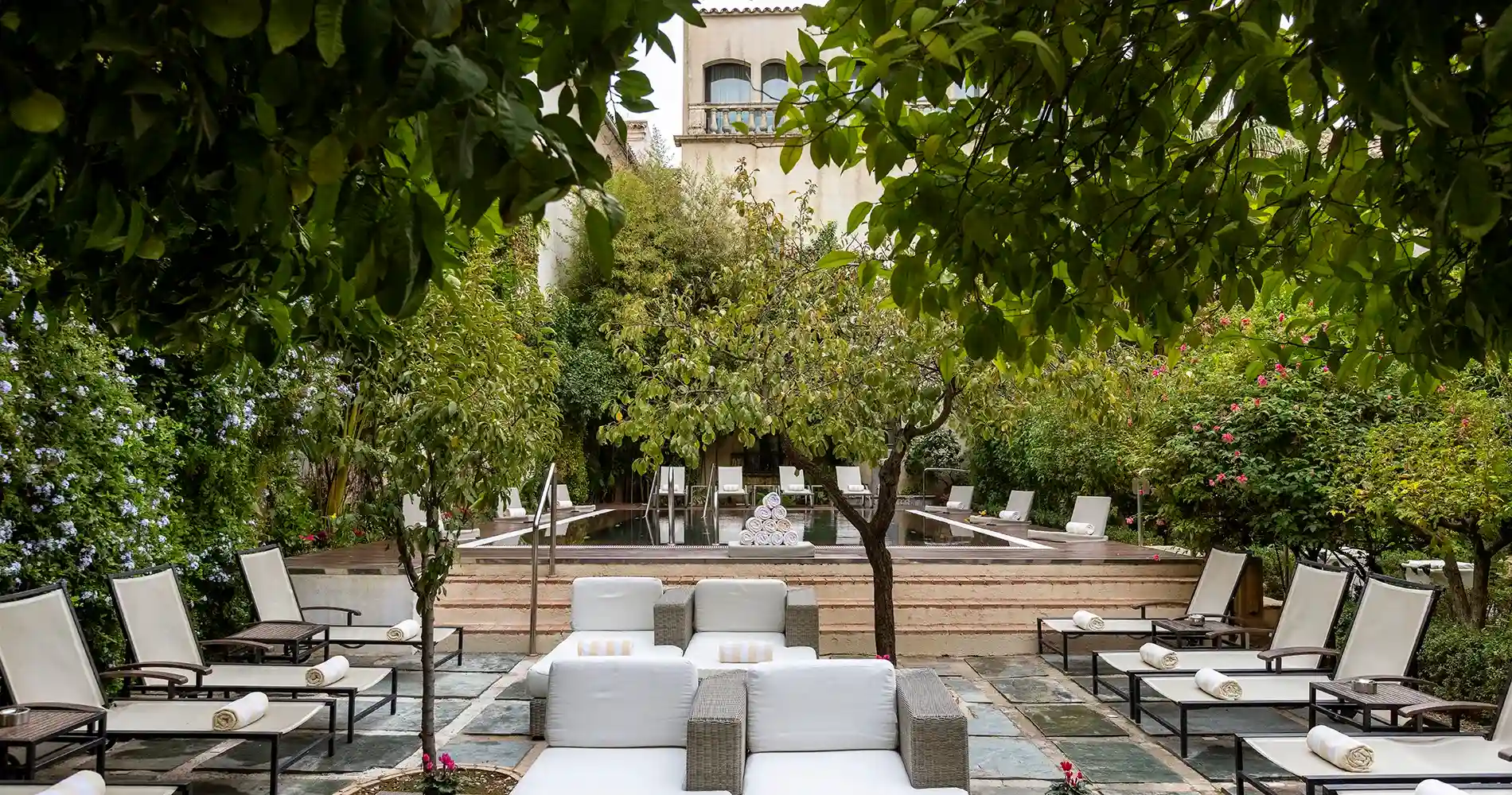 Patio con sof&aacute;s blancos, sillas y piscina rodeada de vegetaci&oacute;n.