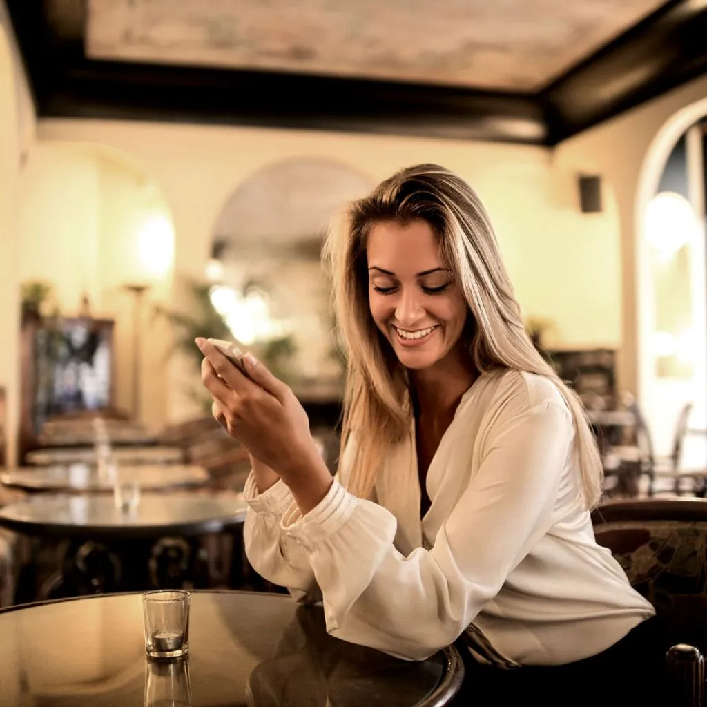 Mujer sonriente mirando su m&oacute;vil en un bar, con luz c&aacute;lida