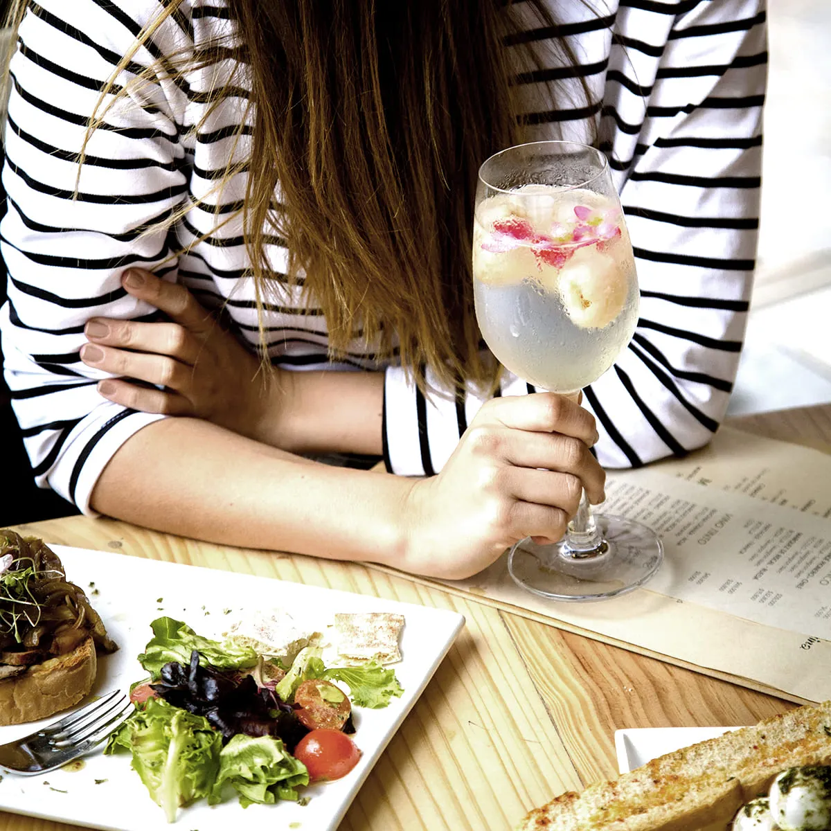 Mujer con blusa a rayas brinda con copa de vino y come en mesa.