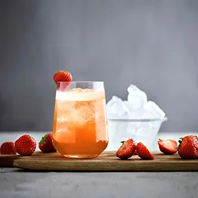 C&oacute;ctel rosado en vaso con hielo, fresas y frambuesa sobre mesa.
