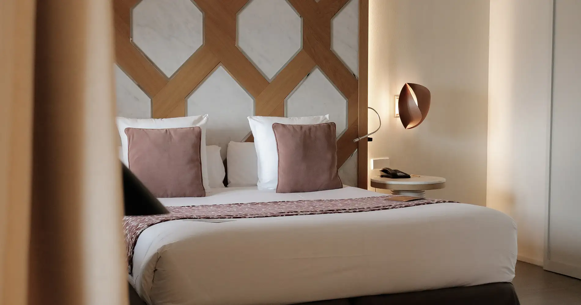 Cama doble con cojines rosados y cabecero geom&eacute;trico de madera, iluminaci&oacute;n c&aacute;lida