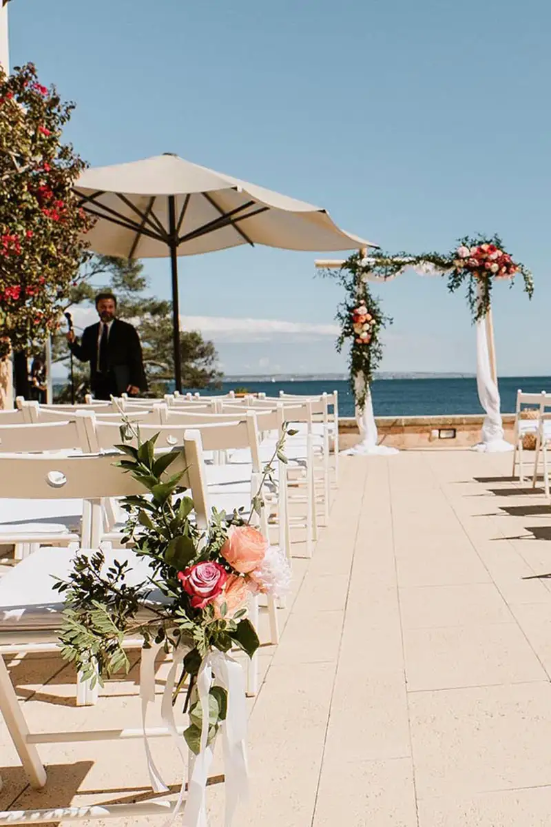 Boda al aire libre junto al mar, sillas blancas y flores decorativas bajo sombrillas