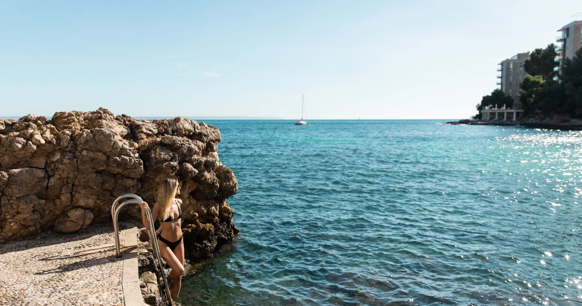 Mujer con bikini en escaleras junto al mar y un velero al fondo