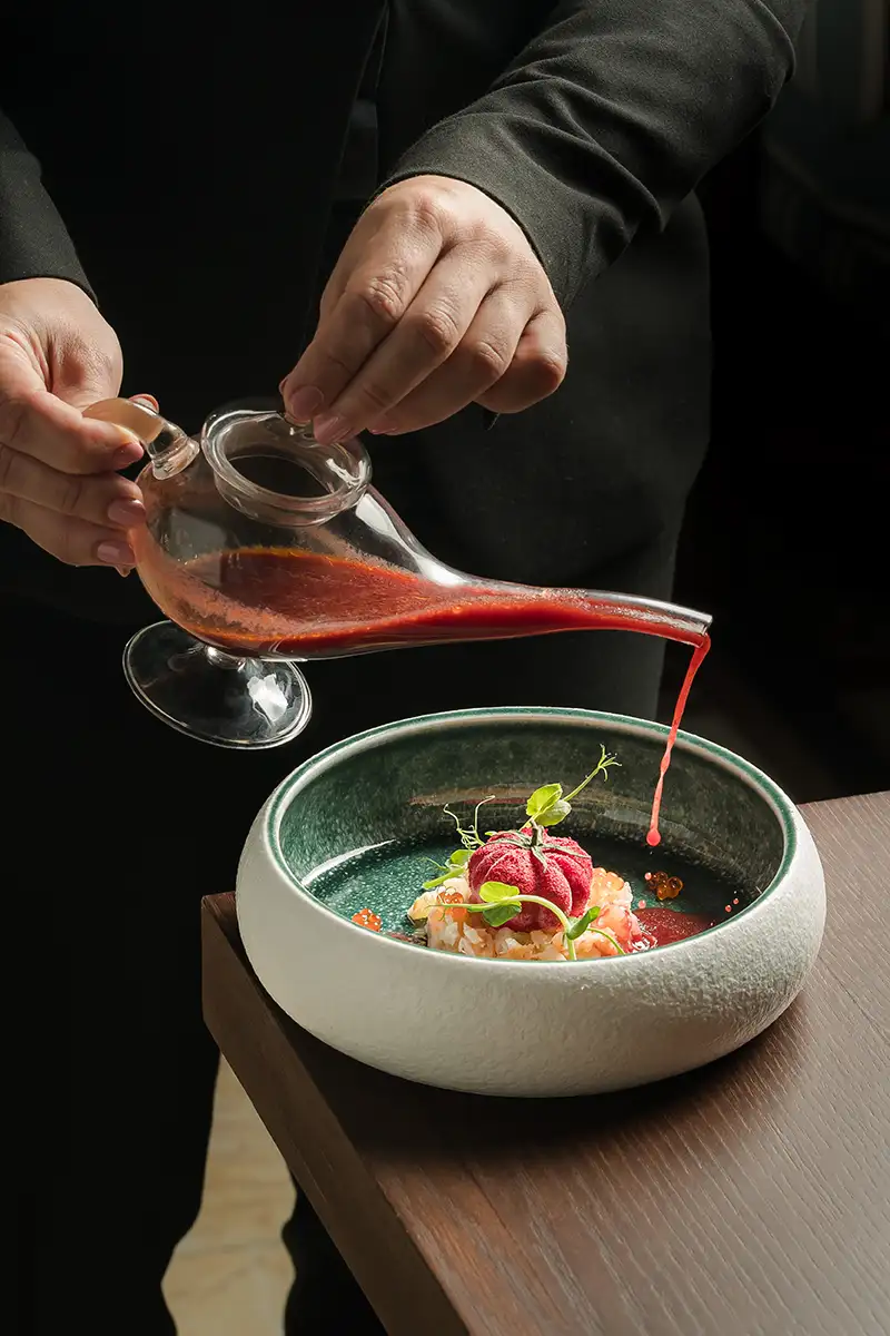 Mano sirve salsa roja desde un recipiente de vidrio sobre un plato gourmet