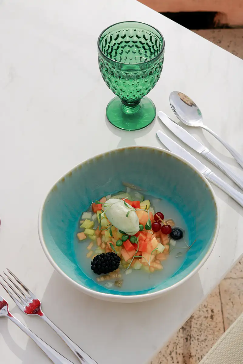 Ensalada servida en plato azul, con copa verde y cubiertos sobre mesa blanca