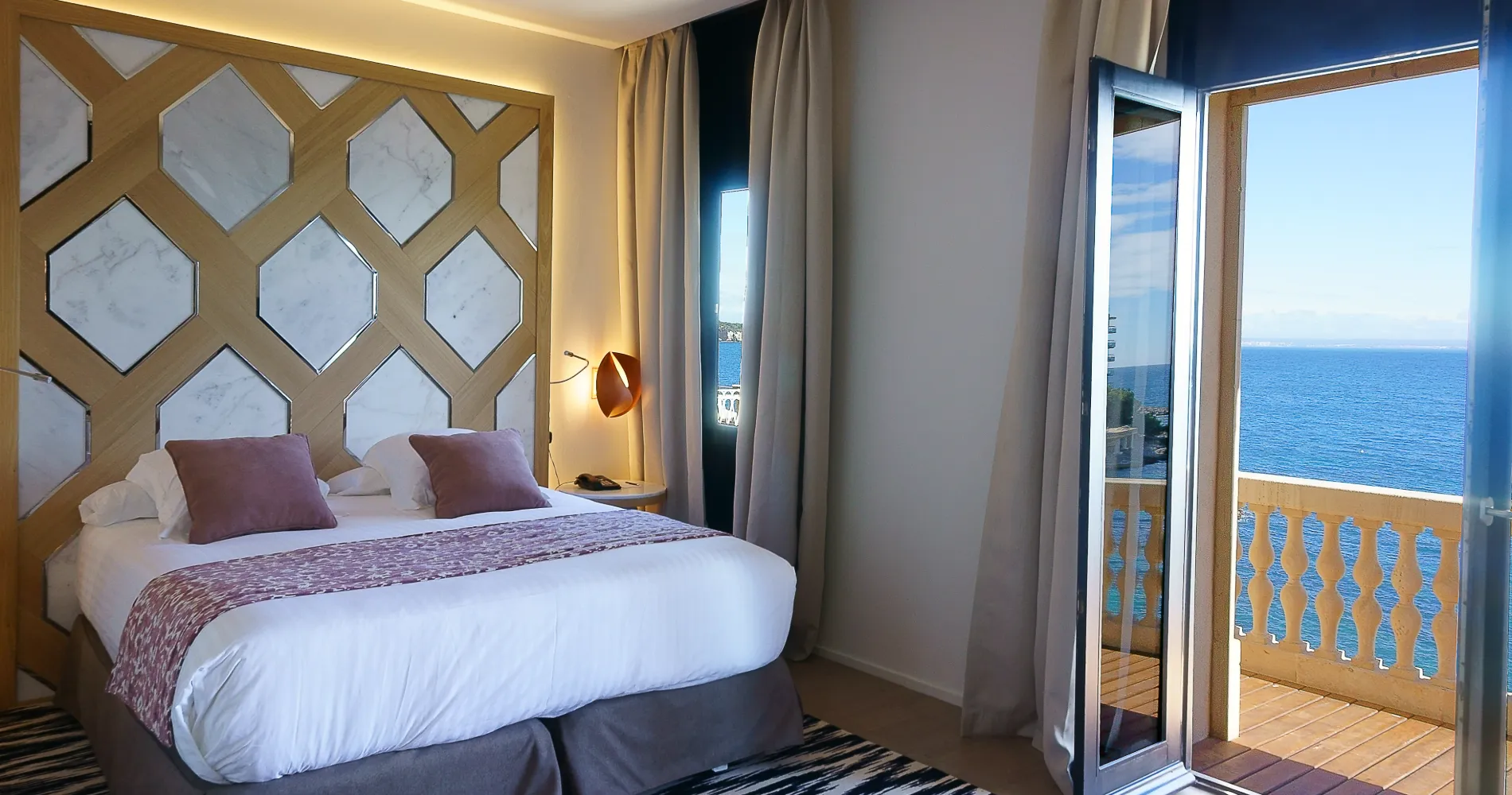 Dormitorio con cama y cortinas, panel hexagonal y vista al mar desde el balc&oacute;n