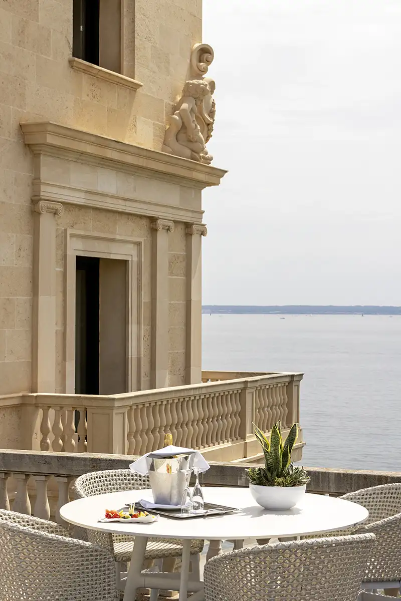 Terraza con mesa, sillas y estatuas sobre el mar, junto a un edificio de piedra
