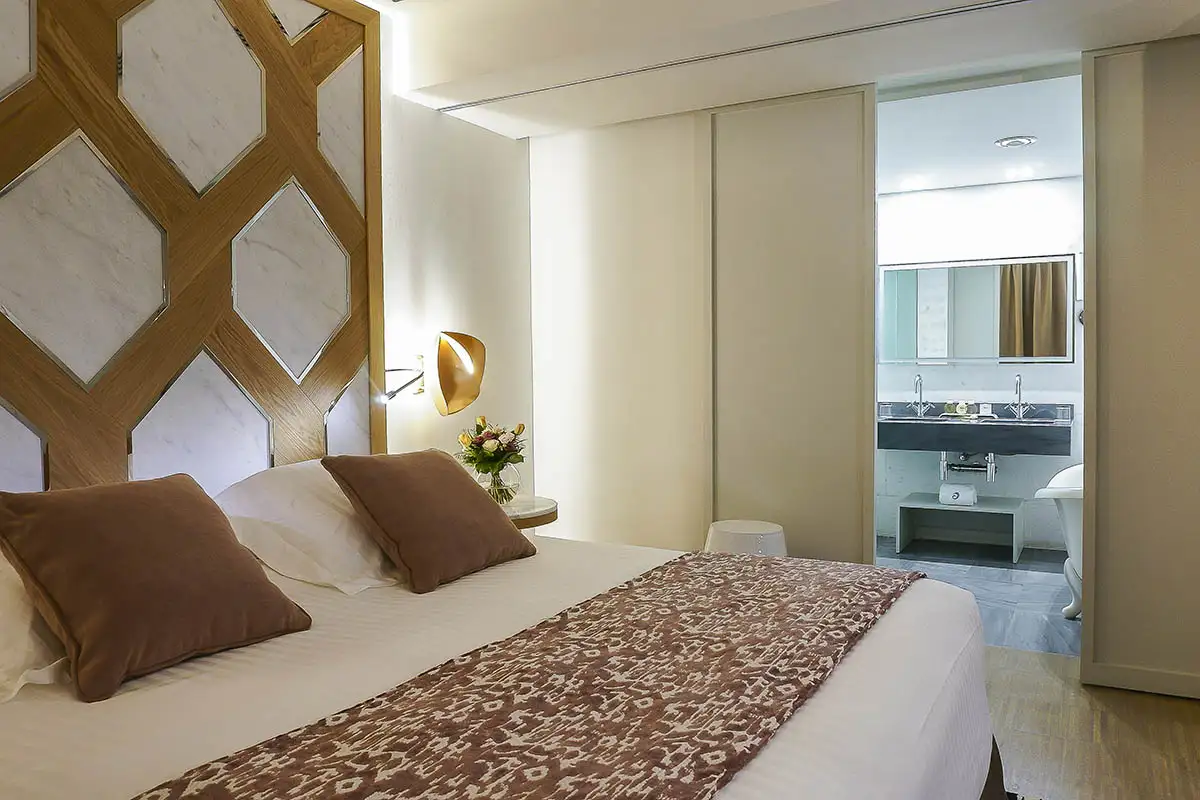 Dormitorio moderno con cama blanca, manta estampada y ba&ntilde;o al fondo