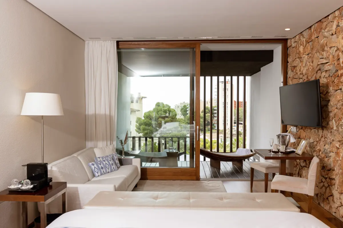 Dormitorio moderno con cama, sof&aacute;, TV y ventana con vista al jard&iacute;n