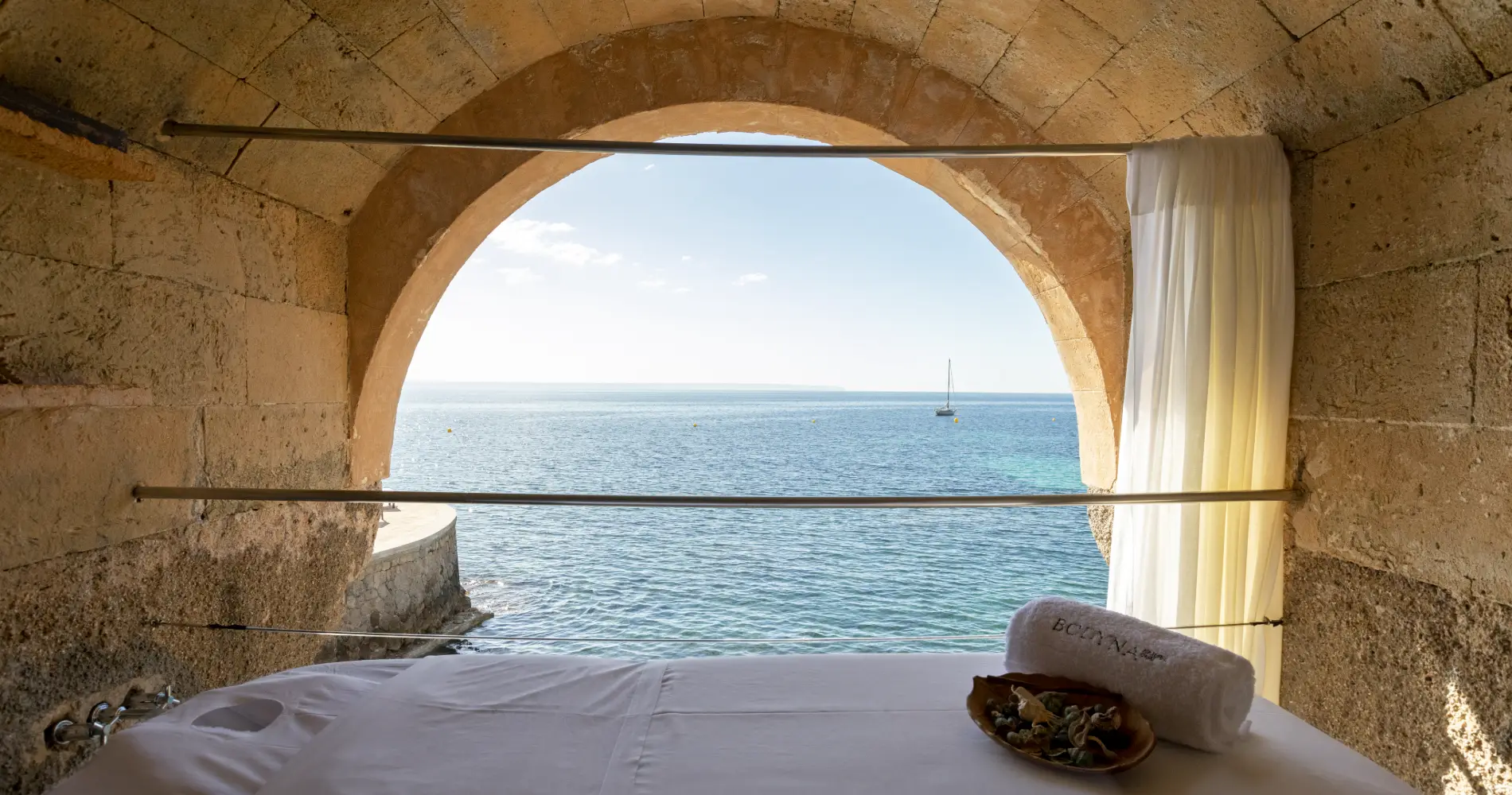 Ventana arqueada con cortina y cama; vista al mar y velero al fondo