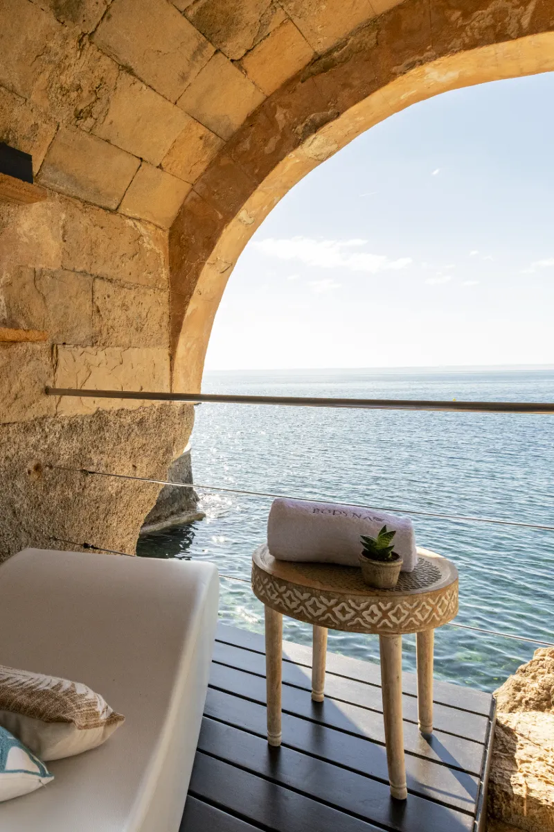 Terraza junto al mar con sill&oacute;n y mesa decorada, bajo un arco de piedra