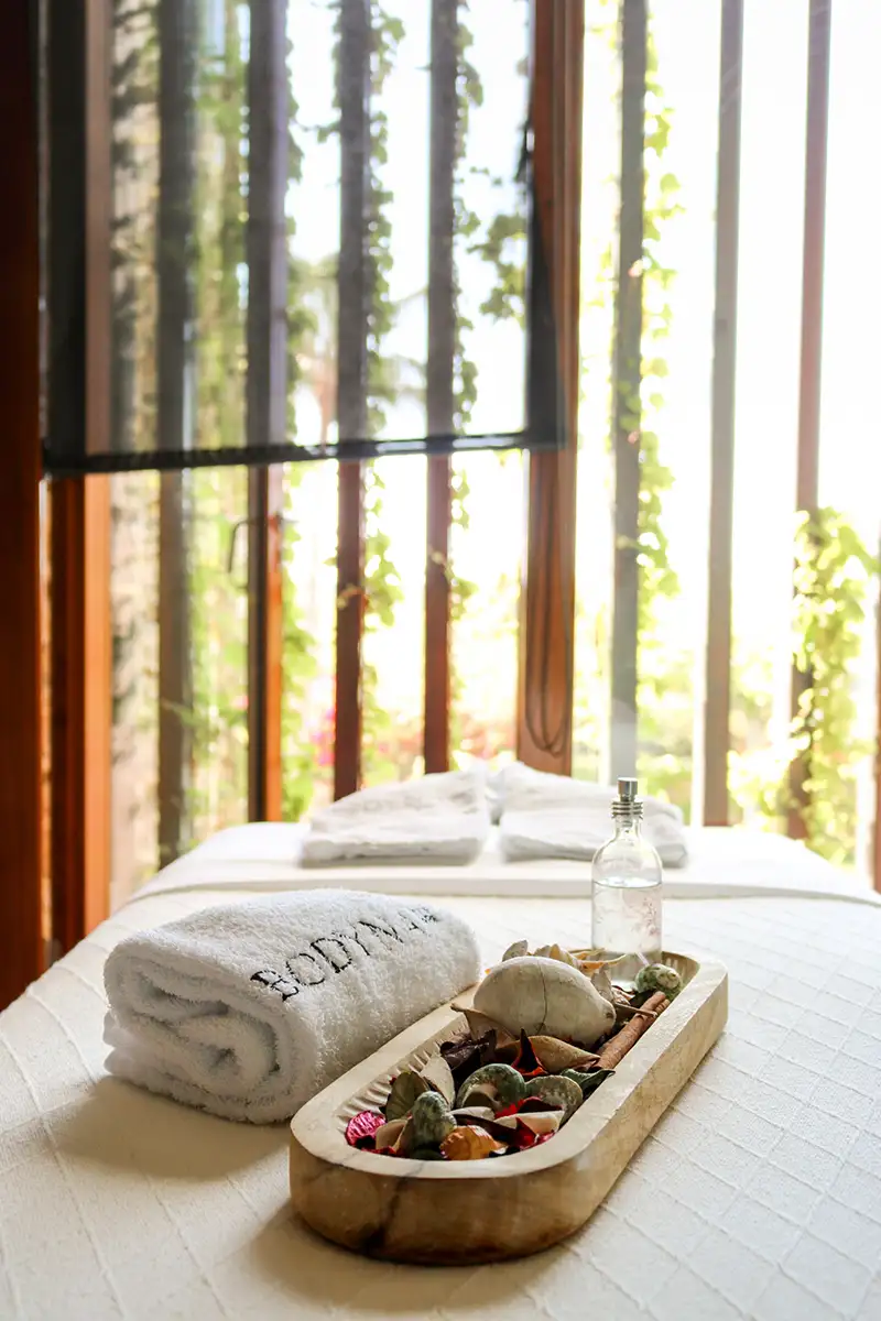 Cama con almohadas y toalla, botella y canastillo junto a ventana con luz.