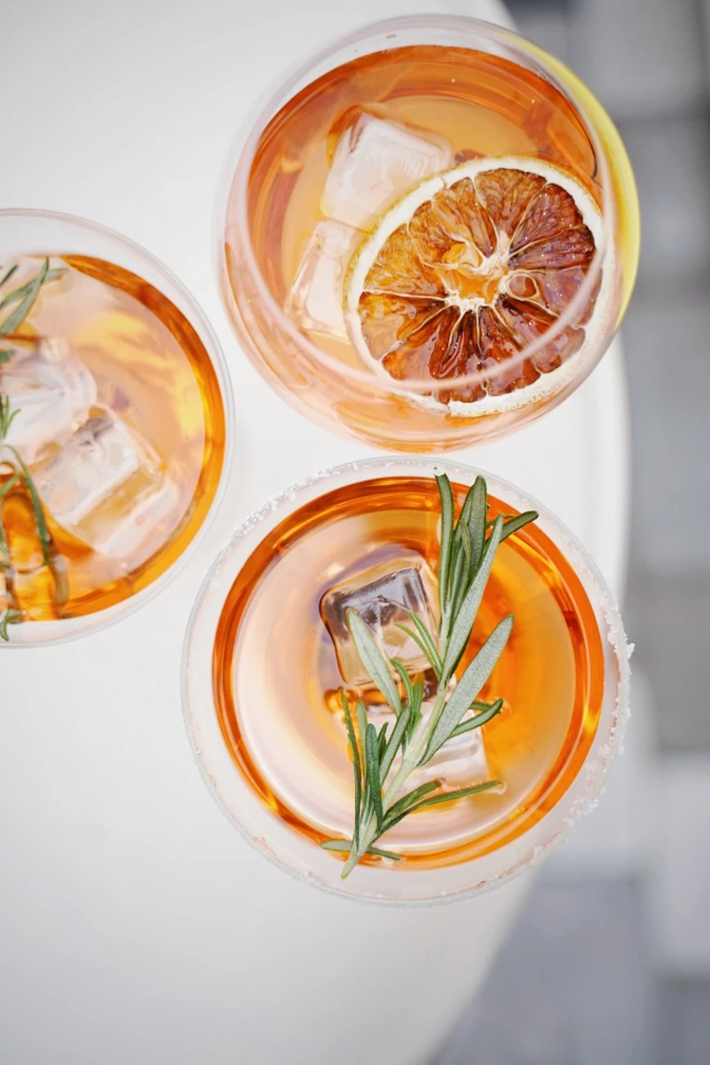Tres c&oacute;cteles de c&iacute;tricos con hielo, rodajas de naranja y romero en vasos.