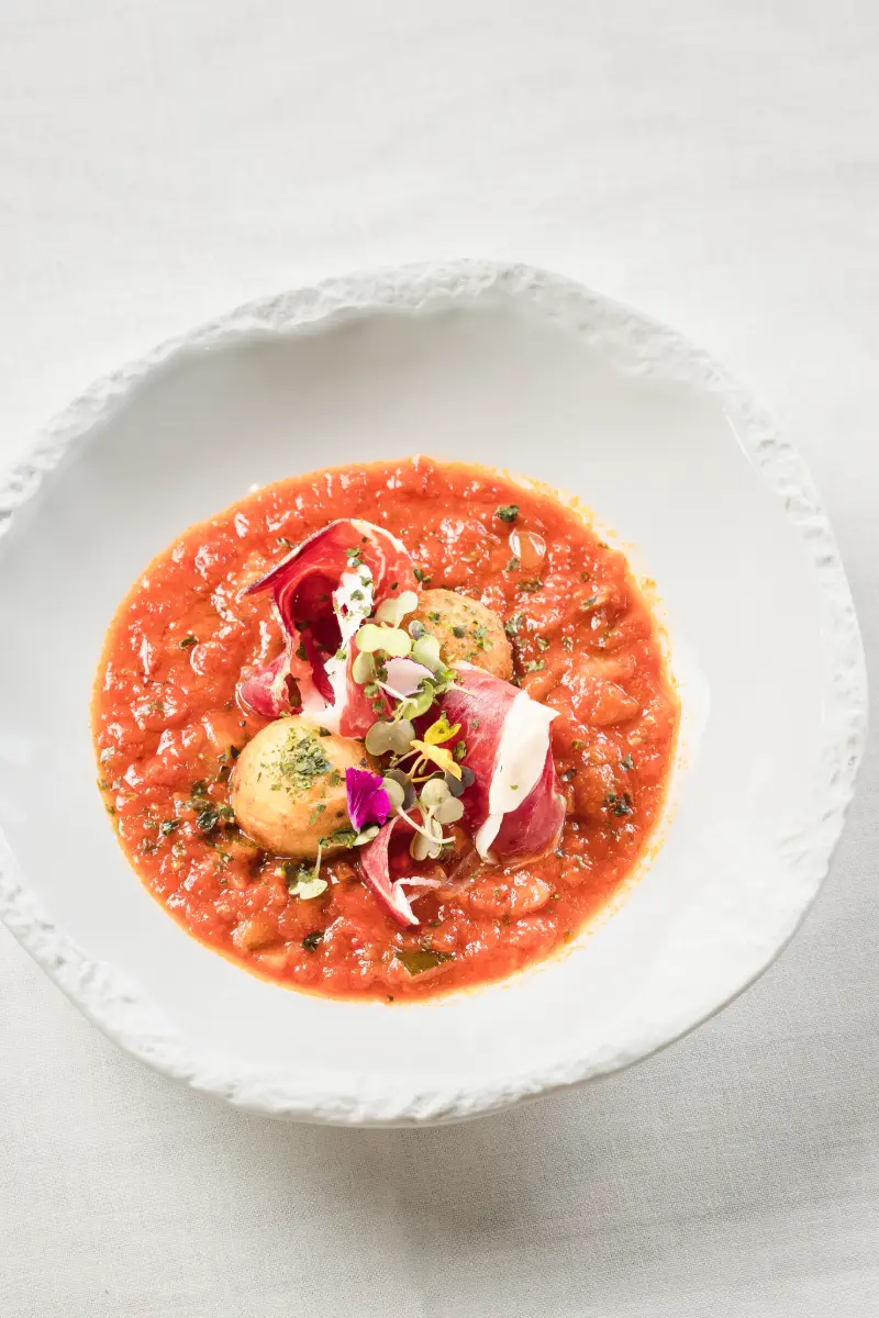 Plato de sopa roja con jam&oacute;n, queso y brotes sobre un plato blanco