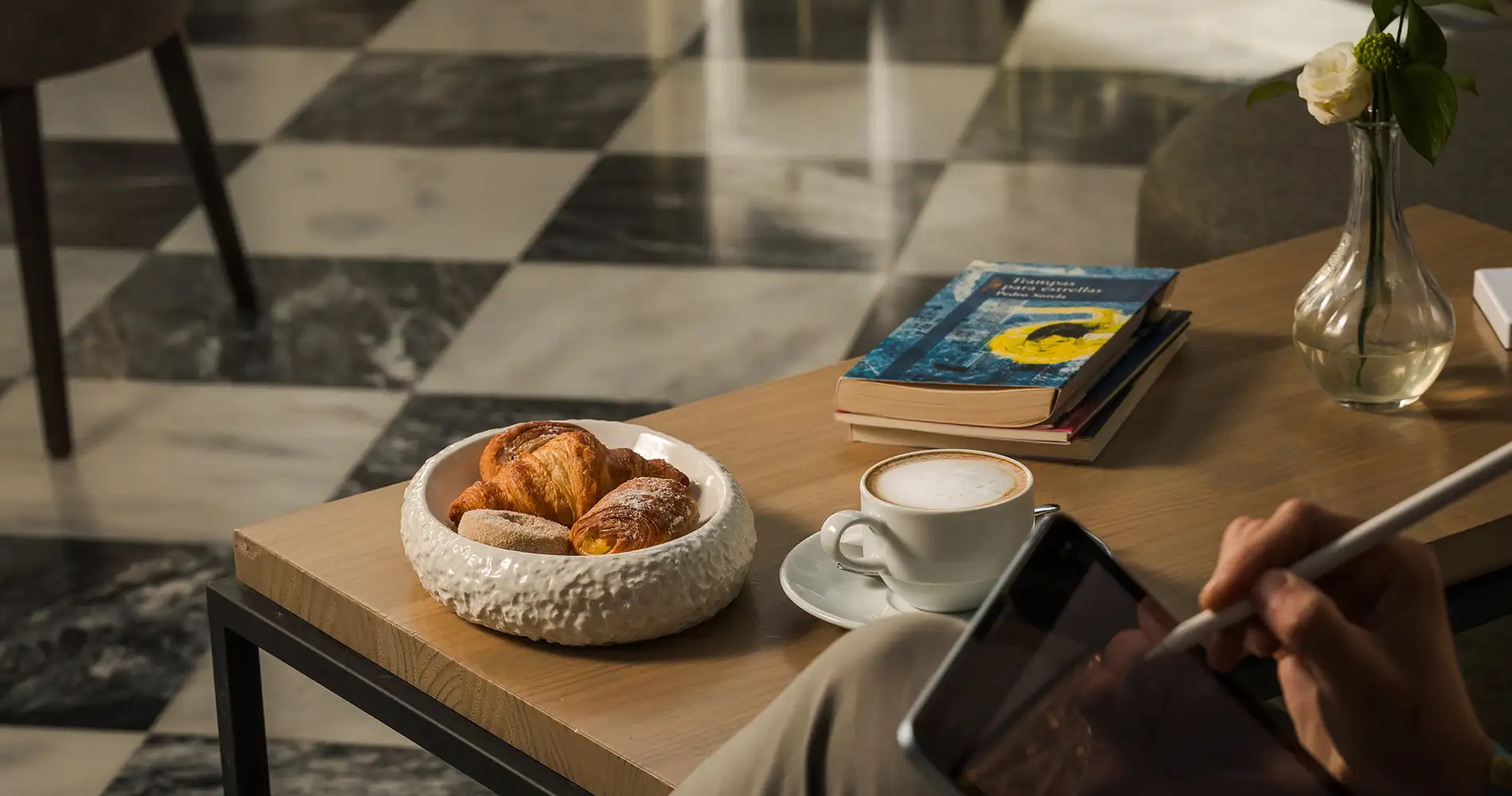 Desayuno con croissants, caf&eacute; y libros en una mesa junto a una mano con m&oacute;vil