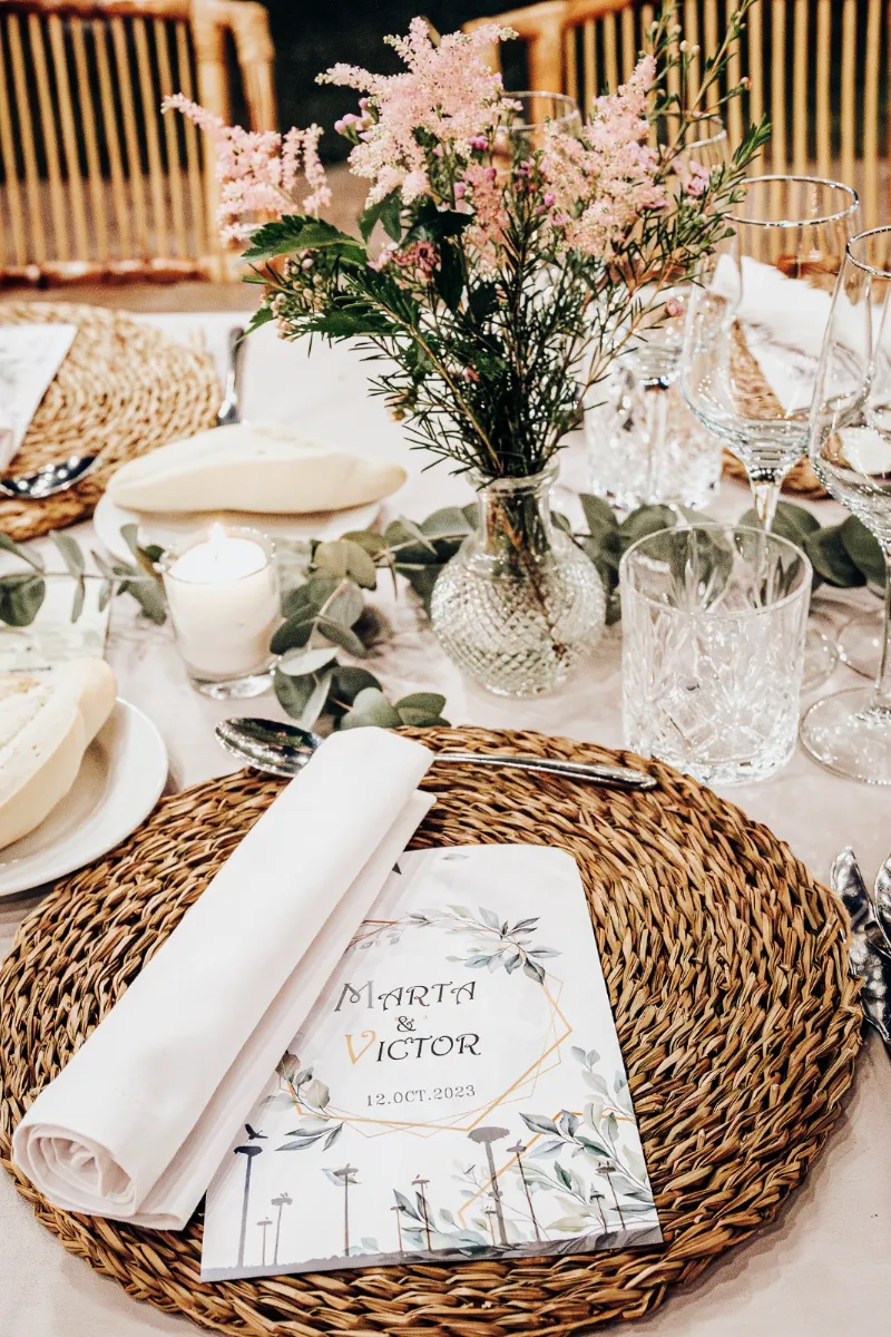Mesa decorada con jarr&oacute;n de flores rosadas, copas y libro de invitaci&oacute;n en mimbre.