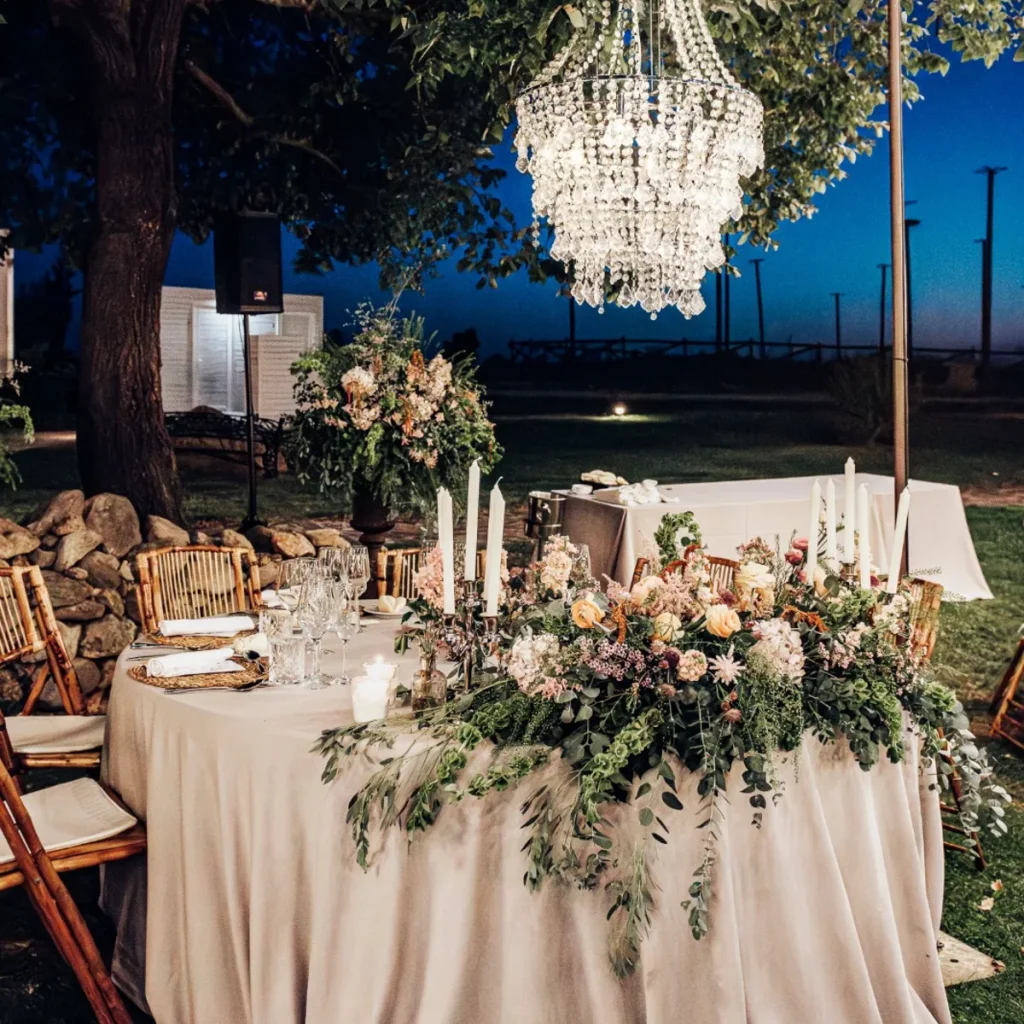 Mesa de banquete al aire libre con flores, velas y l&aacute;mpara de cristal colgante