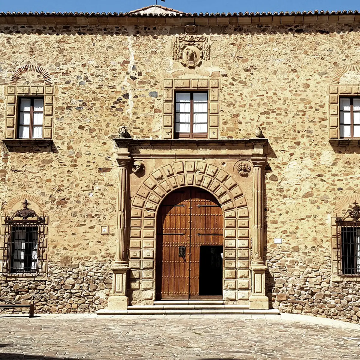 Fachada de piedra con gran puerta de madera y arco tallado, bajo cielo azul