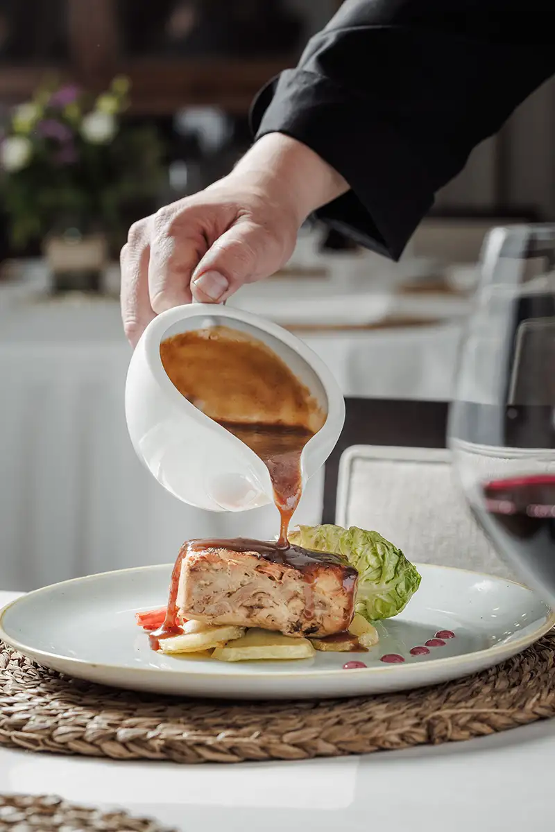 Camarero sirve salsa desde un cazo sobre un plato con carne y ensalada