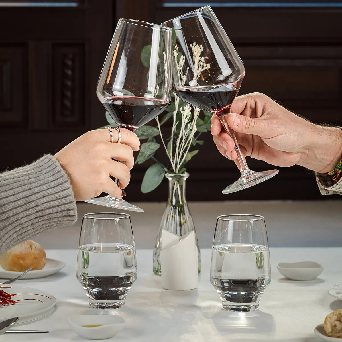 Dos manos brindando con copas de vino tinto en mesa con comida y flores