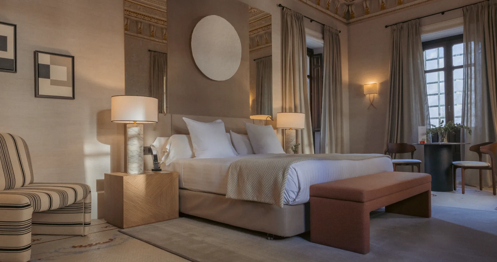 Dormitorio con cama, sillones laterales, l&aacute;mparas y cortinas junto a ventanas