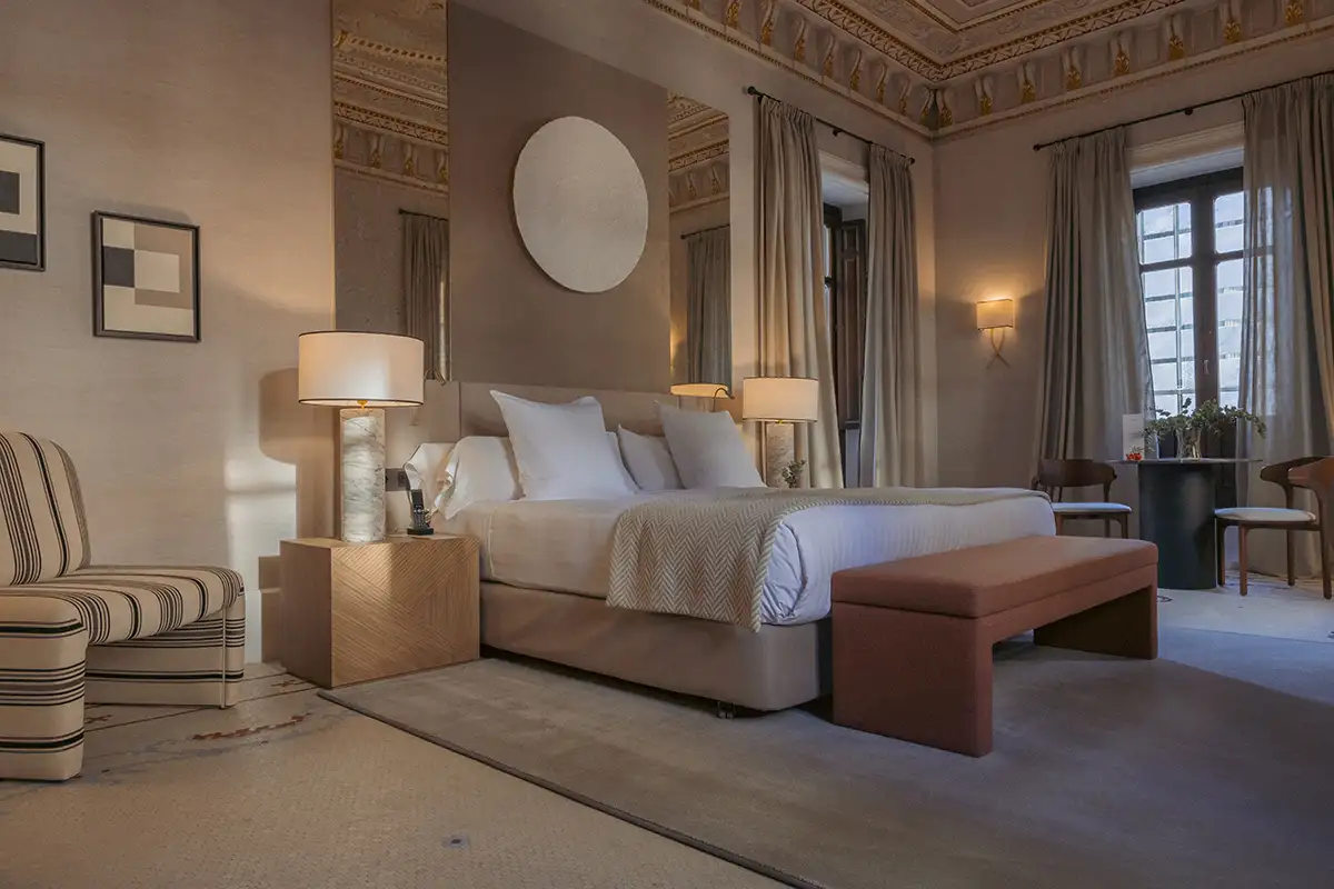Dormitorio elegante con cama, butacas a rayas, l&aacute;mparas y cortinas largas