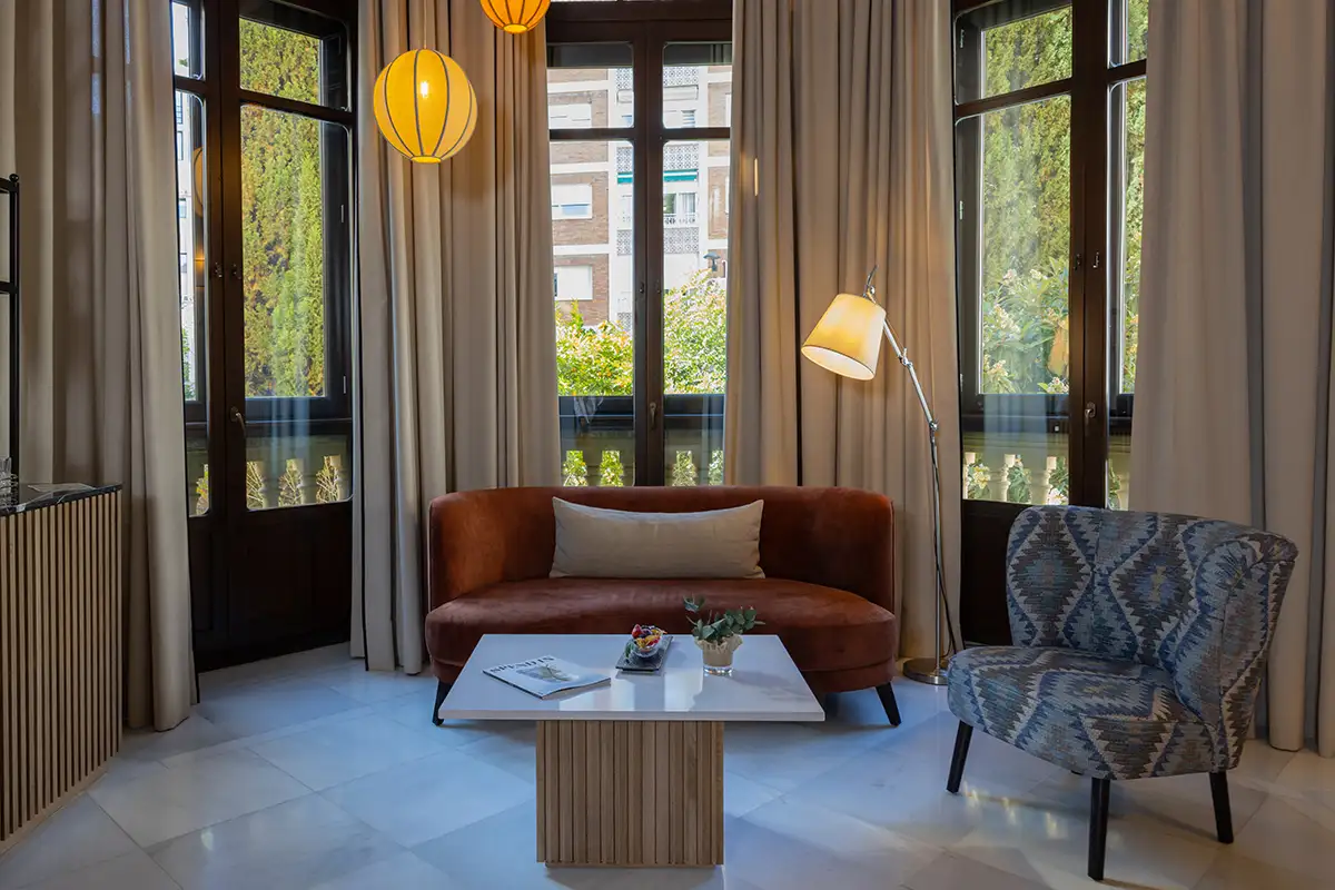 Sala luminosa con sof&aacute; marr&oacute;n, mesa de madera y sill&oacute;n junto a ventanales