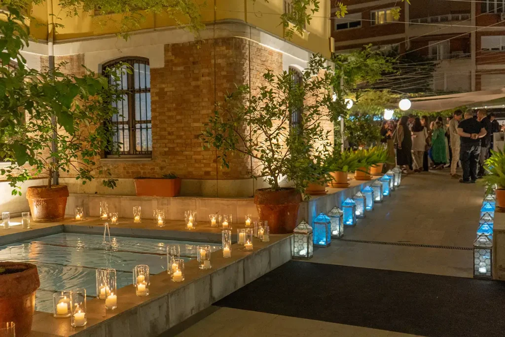 Patio con fuente iluminada, velas y plantas; gente esperando en el pasillo