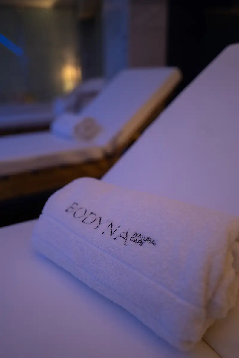 Manos y toalla blanca con bordado &ldquo;F.C. &hellip; Natura&rdquo; en spa, vista cercana.