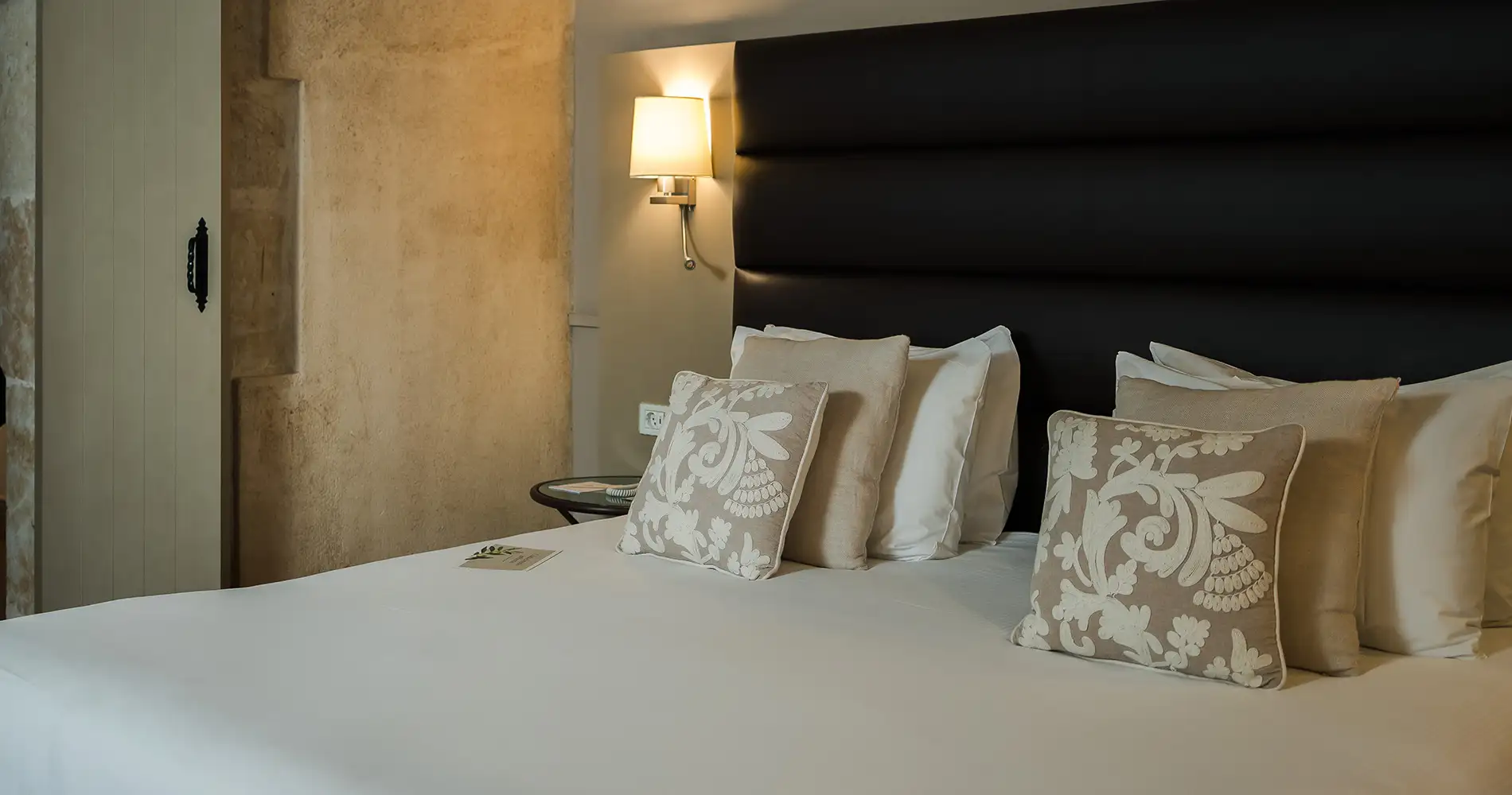 Cama con almohadas decorativas, cabecero oscuro y l&aacute;mpara de pared iluminada