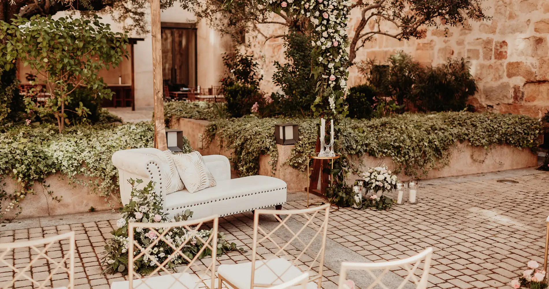 Sof&aacute; blanco con cojines, flores y guirnalda en patio de piedra con jard&iacute;n