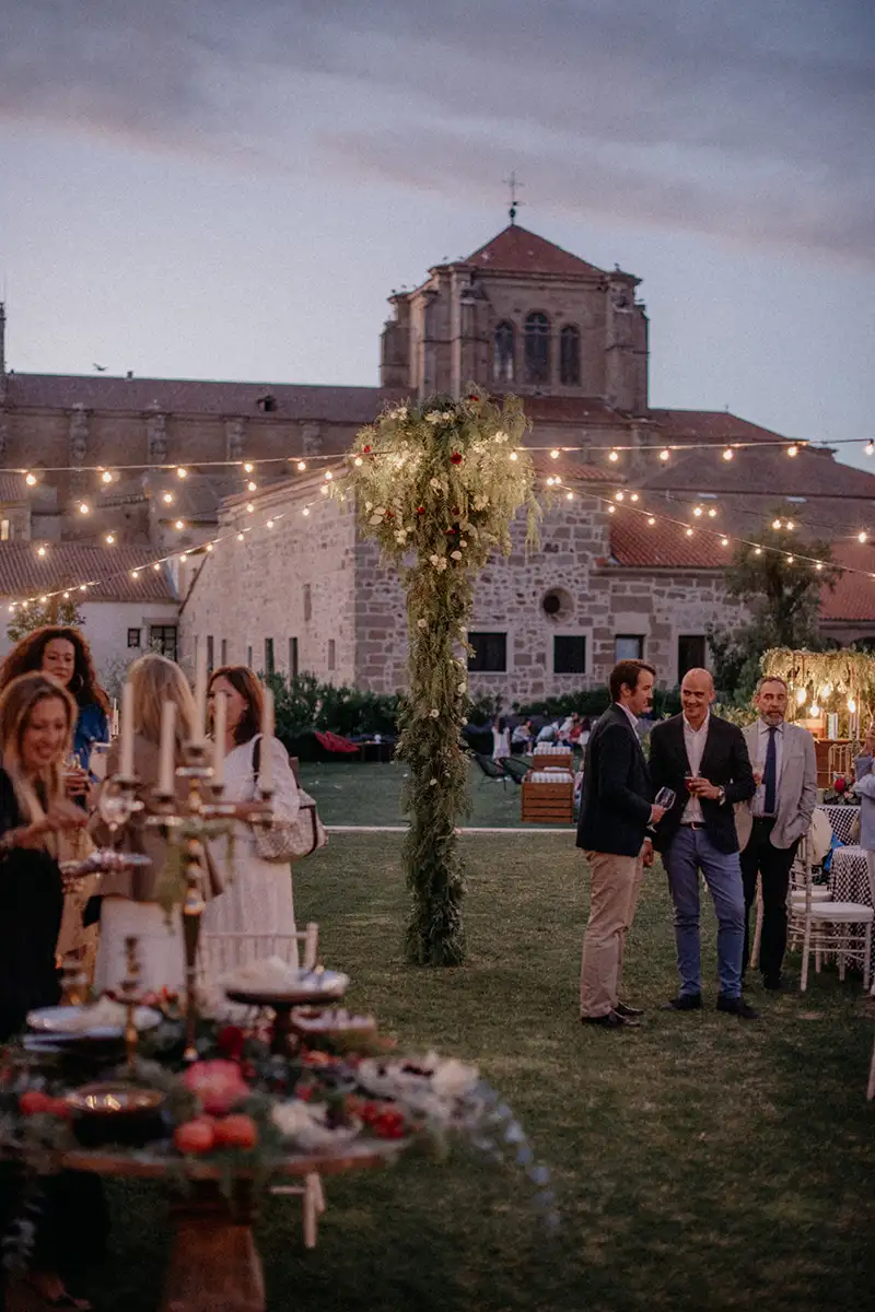 Boda en jard&iacute;n con mesas decoradas y grupo de invitados frente a una iglesia.