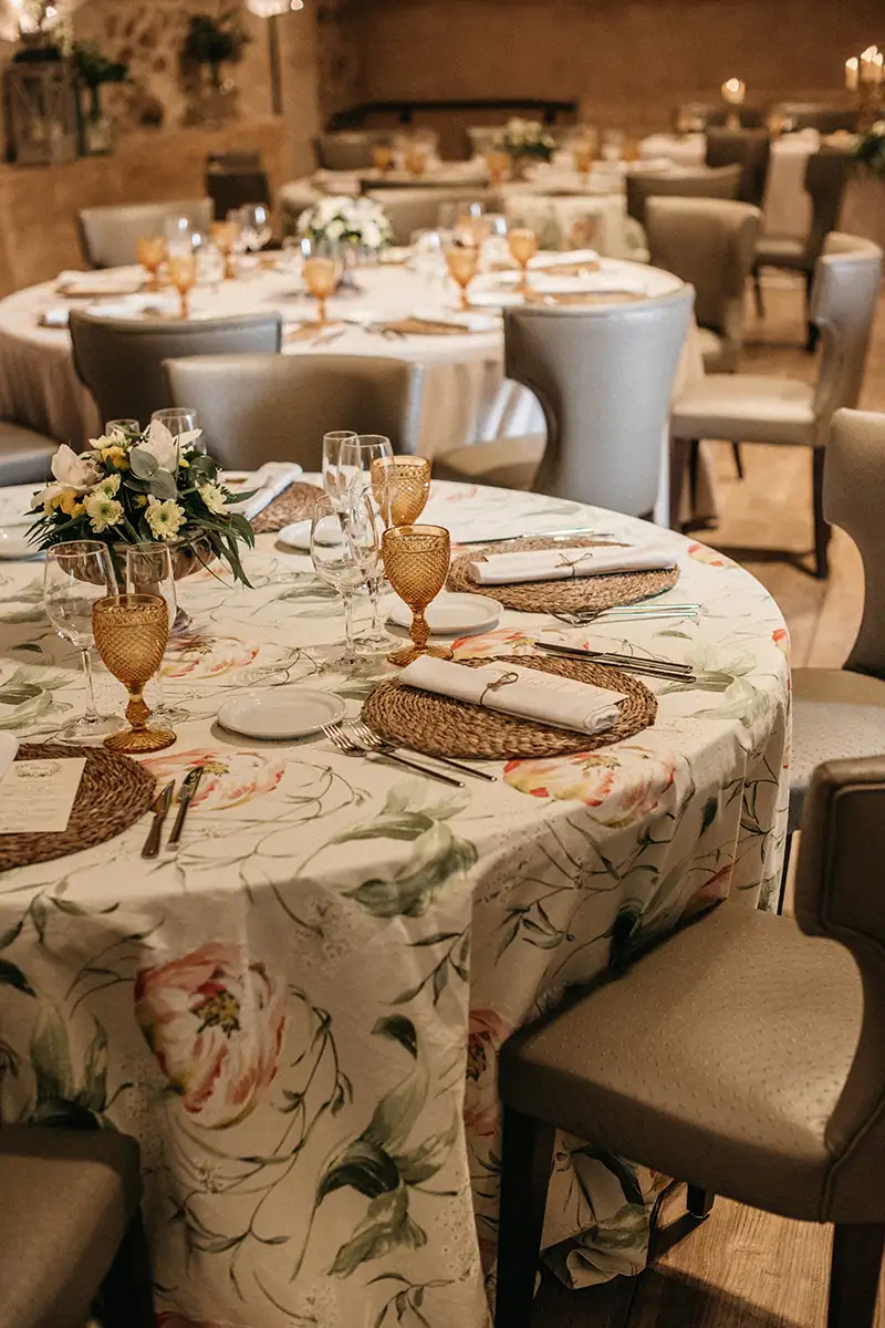 Mesa de banquete con mantel floral, copas, cubiertos y flores en un sal&oacute;n.