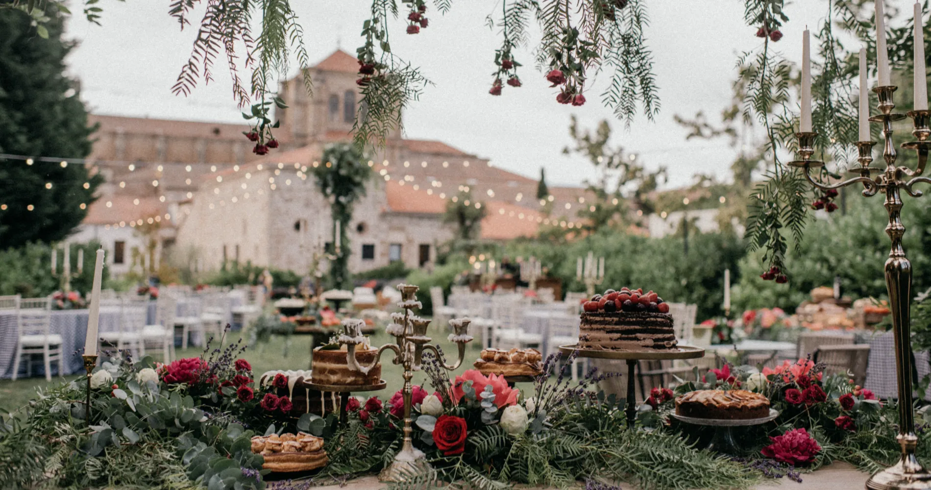 Mesa de postres con flores y velas, fondo de edificio y candelabros decorativos.