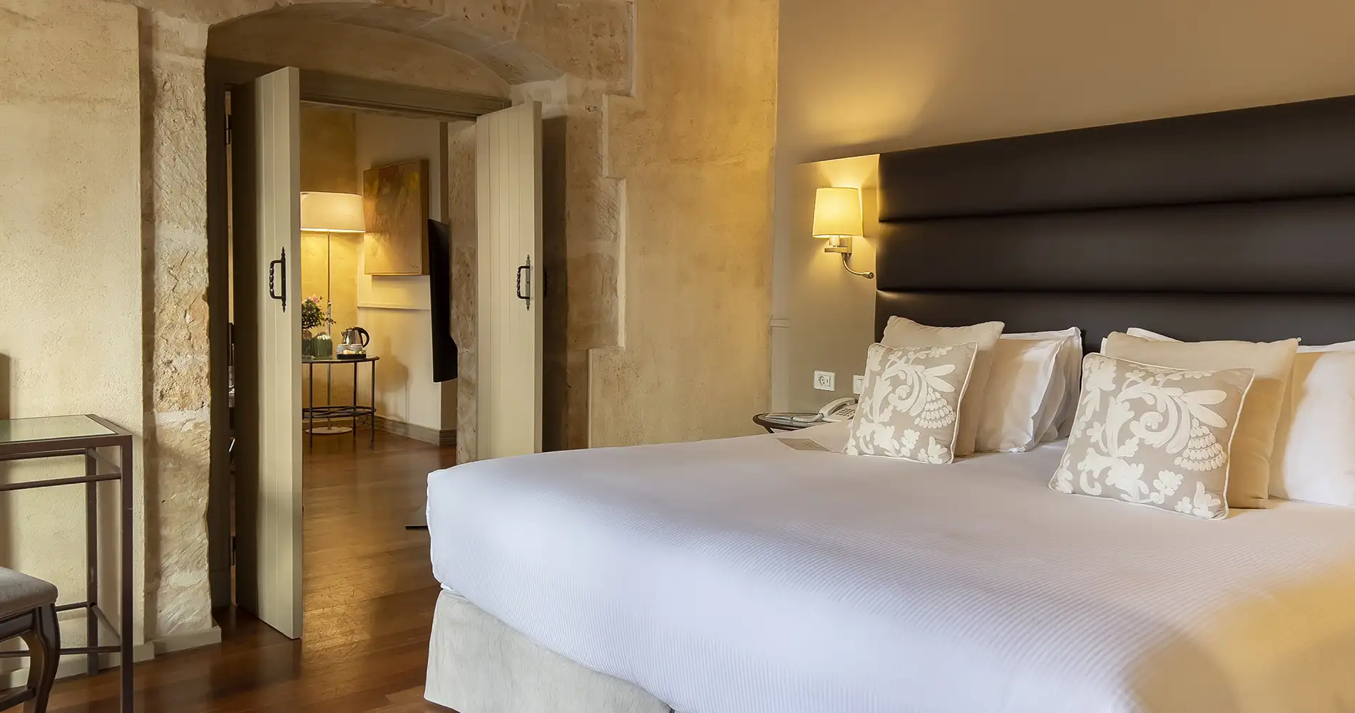 Habitaci&oacute;n de hotel con cama blanca, l&aacute;mparas y pared de piedra, vista interior