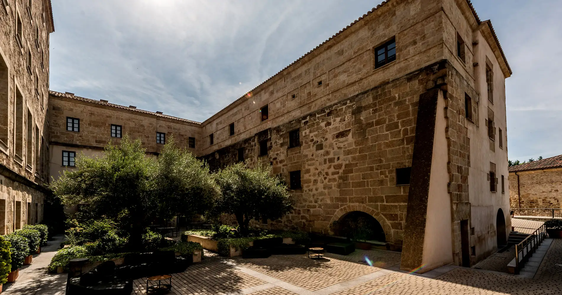 Patio de piedra con arcos, bancos y &aacute;rboles bajo cielo nublado