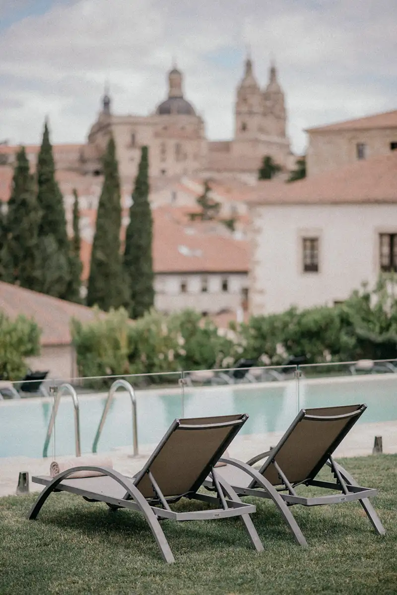 Dos tumbonas junto a una piscina, con el horizonte de edificios hist&oacute;ricos al fondo